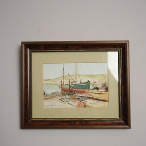 May include: Framed watercolor painting of a boat in a harbor. The boat is red and green, with masts and rigging. The painting is set in a brown wooden frame with a gold inner border. The background shows a shoreline and hills.