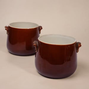 May include: Two matching ceramic pots with a glossy dark brown exterior glaze and a smooth cream-coloured interior. Each pot features a rounded body, a wide circular opening, and two small, decorative handles on opposite sides of the rim.
