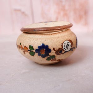 May include: A small, round, speckled beige ceramic jar with a matching lid. The exterior features hand-painted blue flowers, green leaves, and orange accents. A circular white sticker on the side displays the text HECHO EN MEXICO.