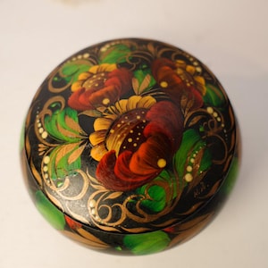 May include: A round, black lacquered box with a floral design. The lid features large, stylized flowers in shades of red, yellow, and orange, with green leaves and gold accents. The box is a decorative item.