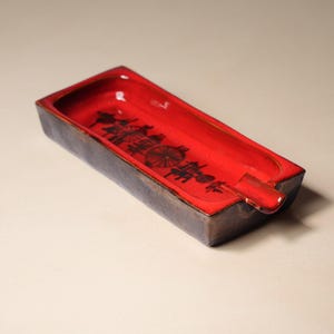 May include: A rectangular ceramic ashtray featuring a glossy, vibrant red interior basin with a black graphic print of stylized wheels and geometric patterns. The exterior sides have a dark, textured, matte finish with a single notch.