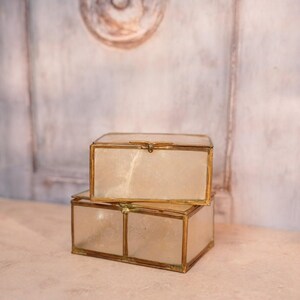 Puede incluir: Dos cajas rectangulares de cristal con marcos de metal color latón están apiladas. Las cajas tienen un interior nacarado, blanquecino. El fondo es una pared clara y texturizada.