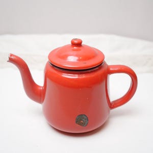 Könnte beinhalten: Kleine rote Emaille-Teekanne mit passendem Deckel und gebogenem Ausguss. Die Teekanne hat einen abgerundeten Körper, einen schwarzen Rand und einen Griff. Auf der Seite ist ein kleines, kreisförmiges Design zu sehen.