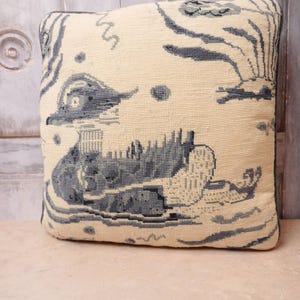 May include: Square cushion with a cream backdrop and a blue and white landscape design. The cushion has a detailed, textured pattern, with a soft, plush appearance. The design includes trees, water, and architectural structures, creating a scenic view.