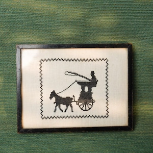May include: Framed black and white cross-stitch depicting a horse-drawn carriage with a driver. The artwork is set against a white background with a black zigzag border, encased in a dark wooden frame. The piece is displayed on a green textured surface.