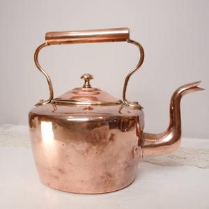 Könnte beinhalten: Eine Vintage-Kupferkanne mit gebogenem Ausguss und Klappdeckel. Die Kanne hat einen abgerundeten Körper, einen Griff und einen kleinen Knopf auf dem Deckel. Das Kupfer hat eine warme, rötlich-braune Farbe.