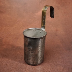 Op de afbeelding: Een cilindrische metalen maatbeker met een verweerde zilveren afwerking en een hoge, gebogen handgreep in messingkleur. Het buitenoppervlak heeft vage reliëftekst en een versleten textuur, terwijl de binnenkant glad en licht van kleur is.