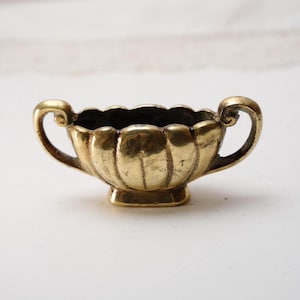 Puede incluir: Una pequeña taza de latón antiguo con dos asas curvas. La taza tiene un borde festoneado y un cuerpo texturizado y acanalado. La base es cuadrada. El interior es oscuro, contrastando con el exterior dorado brillante. Una pieza decorativa.