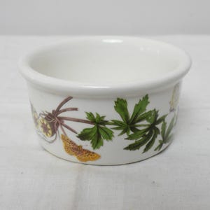 Op de afbeelding: Een kleine, witte keramische kom met een afgeronde rand. De kom is versierd met groene bladeren en een vlinderontwerp in tinten geel, bruin en groen. De kom is waarschijnlijk bedoeld om te serveren of tentoon te stellen.