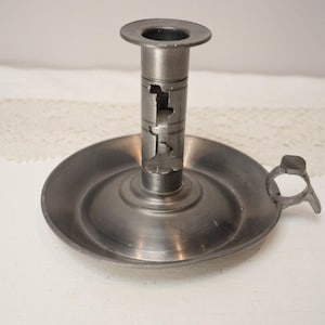 May include: A pewter candlestick holder with a circular base and a central column. The column features cut-out designs and a top ring for a candle. A small handle is attached to the side of the base. The candlestick is made of metal.