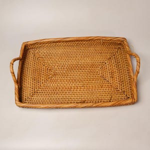 May include: A rectangular woven rattan tray featuring a light brown natural finish, a tight grid weave pattern on the base, and two integrated curved handles on the shorter sides. The tray has a raised border and a consistent woven texture.