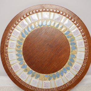 Pode incluir: Bandeja redonda de madeira com borda de mosaico. O mosaico apresenta um padrão de azulejos brancos, azuis e castanhos. O centro da bandeja é de madeira maciça castanha escura. A bandeja tem cerca de 30 cm de diâmetro.