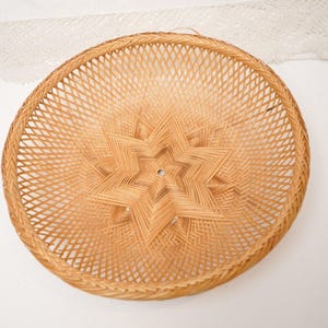 May include: Circular woven basket featuring a star-shaped design at its centre. Constructed from light brown wicker, the basket showcases a woven pattern. The starburst design is a key element, enhancing the visual appeal of the item.