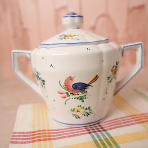 May include: A white ceramic sugar bowl with a lid, featuring hand-painted motifs of a small bird with a red head and blue wings, surrounded by green leaves and yellow flowers. The bowl has two angular handles and blue trim along the edges.