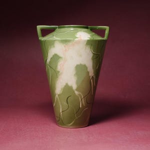May include: A sage green ceramic vase with two handles at the top rim. The body features a large, central white cloud-like pattern with soft pink accents and thin, dark green wavy lines that extend downwards across the surface of the vessel.