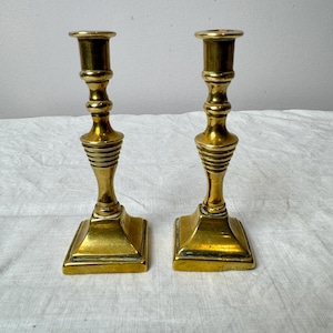 Peut inclure: Deux bougeoirs anciens en laiton. Chaque bougeoir a une base carrée, une tige détaillée avec des rainures horizontales et un sommet en forme de coupe. Les bougeoirs sont de couleur or chaud et semblent être en métal. Ils sont posés sur une surface blanche.