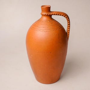 May include: A terracotta-coloured ceramic jug with a rounded body, a narrow neck, and a single side handle. The surface features horizontal wavy line engravings around the shoulder and a notched, textured pattern along the outer edge of the handle.