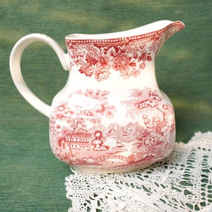 May include: A white ceramic jug with a red floral and landscape pattern. The jug features a curved handle and a spout. The design includes flowers, trees, buildings, and bridges. The jug is placed on a white lace doily.
