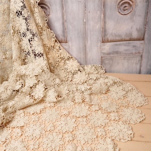 May include: A cream-colored, openwork lace textile with a floral pattern. The lace is draped over a light-colored wooden surface, with a portion of a weathered, gray door in the background. The edges of the lace have a scalloped trim.