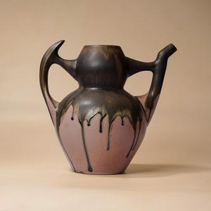 May include: A ceramic pitcher with a bulbous body and two handles. The vessel features a matte mauve base colour with a dark brown glaze dripping from the top rim down the sides. It has a wide opening at the top and a pointed spout on the right side.