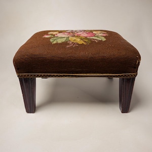 May include: A rectangular footstool featuring a dark brown needlepoint top with a central floral motif in shades of pink, yellow, and green. The stool is supported by four dark wooden legs with a vertical grooved design and a decorative trim.