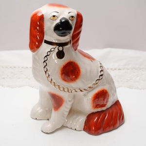 May include: A ceramic dog figurine in a seated position. The dog is primarily white with red accents on its ears, spots, and tail. It has a black nose and a gold-colored chain around its neck. The figurine is a decorative item.