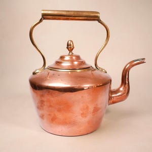 May include: A polished copper tea kettle featuring a rounded body, a curved spout, and a sturdy arched handle. The lid is topped with a small, rounded finial. The metal surface displays a warm, metallic orange-brown tone throughout.