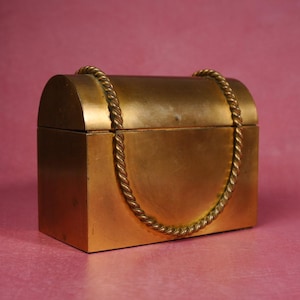May include: A rectangular brass box with a rounded, arched lid. The box features a decorative, twisted rope-style metal handle that extends from the top of the lid down the front face, creating a U-shaped accent against the smooth metal surface.