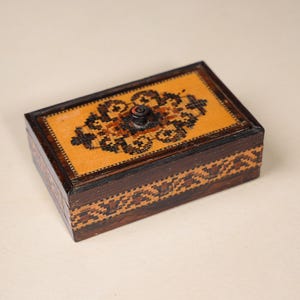 May include: A rectangular wooden box featuring a dark brown finish and a light tan lid. The lid displays a central black and brown geometric pattern with a small black knob. The sides of the box are decorated with a repeating brown and tan pattern.