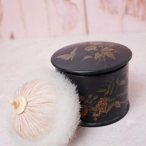 May include: A round black powder jar with a lid featuring gold floral and butterfly illustrations, accompanied by a soft, pale pink powder puff with a white fluffy trim and a small central handle on top of the puff.