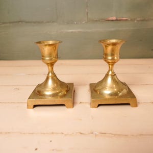 Op de afbeelding: Twee antieke messing kandelaars. Elke houder heeft een vierkante basis, een afgeronde steel en een uitlopende bovenkant. Het messing heeft een warme, gouden tint en de houders staan op een lichtgekleurd houten oppervlak.