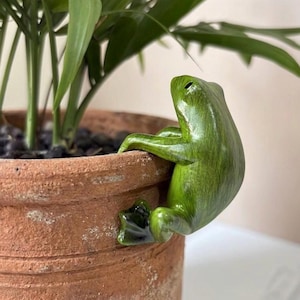 May include: A green frog ornament clinging to the rim of a terracotta plant pot. The frog has a glossy finish and is positioned near a green plant. The pot contains dark soil and is partially filled with a green plant.