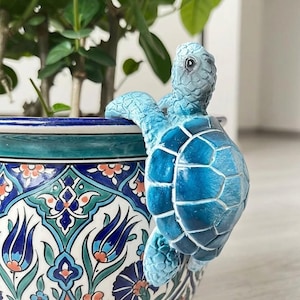 May include: A blue ceramic turtle ornament clinging to the rim of a patterned ceramic plant pot. The turtle has a textured shell and is painted in shades of blue. The pot features a floral design with blue, white, and orange accents.