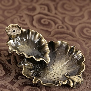 May include: A metallic brass-toned incense holder shaped like two overlapping lotus leaves. The upper leaf features a small lotus seed pod attachment, while the lower leaf includes a small frog figure resting on the edge with detailed veining.