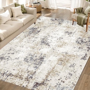 Puede incluir: Una alfombra rectangular grande con un diseño abstracto. La alfombra presenta un fondo crema con parches grises, marrones y azul oscuro. Está colocada sobre un suelo de madera, con un sofá y un sillón a la vista.