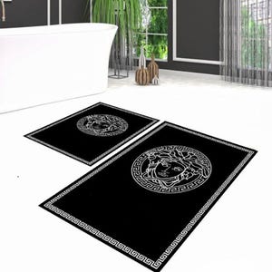 May include: Black bathroom rug set featuring a white Medusa head design. The set includes a rectangular rug and a smaller square rug. The rugs have a white border with a Greek key pattern. The bathroom has a white tub and dark gray walls.