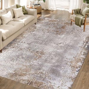 Op de afbeelding: Een rechthoekig vloerkleed met een distressed, abstract ontwerp. Het vloerkleed heeft een mix van grijze, witte en bruine tinten, waardoor een textuur ontstaat. Het vloerkleed ligt op een houten vloer, in een woonkamer.