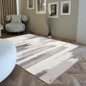 May include: A neutral-toned area rug with a brushstroke design in shades of gray, beige, and white. The rug is placed on a wooden floor, and the room features framed artwork, a patterned chair, and a vase with decorative branches.