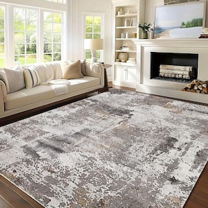 May include: A large area rug with an abstract design in shades of gray, white, and tan. The rug has a distressed, painterly appearance, adding a modern touch to the living room setting. The rug is placed on a hardwood floor.