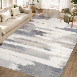 May include: A large rectangular area rug with a watercolor-like design. The rug features broad, horizontal strokes of white, gray, and beige, creating a soft, abstract pattern. The rug is placed in a living room setting.