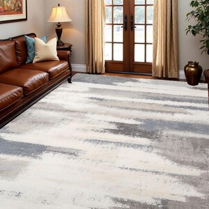 May include: A large area rug with a modern abstract design. The rug features a pattern of horizontal brushstrokes in shades of white, gray, and light beige. The rug is placed on a hardwood floor in a living room setting, with a brown leather sofa visible.