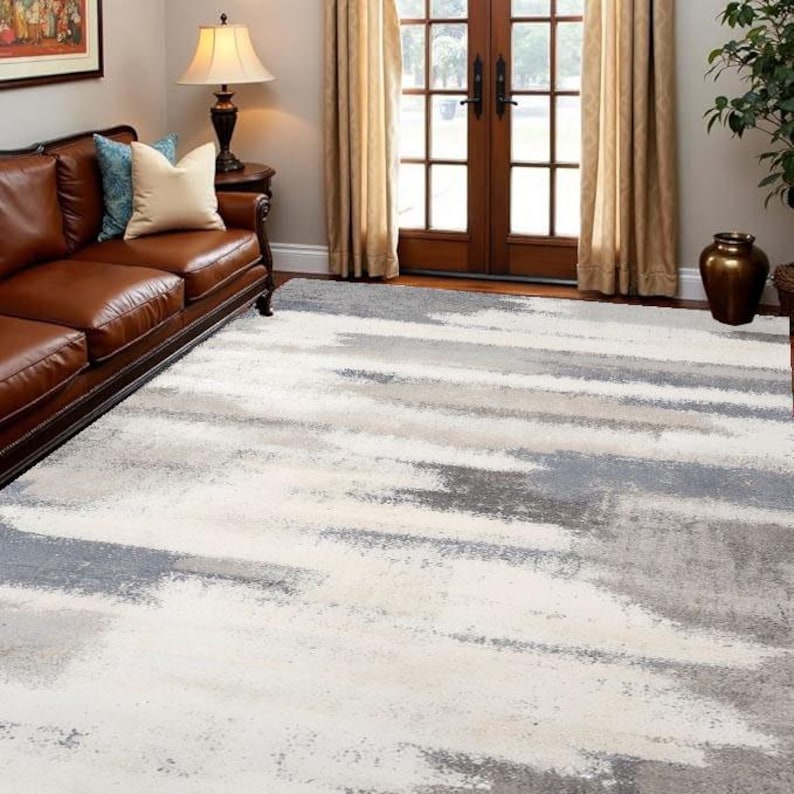 May include: A large area rug with a modern abstract design. The rug features a pattern of horizontal brushstrokes in shades of white, gray, and light beige. The rug is placed on a hardwood floor in a living room setting, with a brown leather sofa visible.