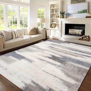 May include: A large area rug with a modern abstract design. The rug features a pattern of broad, irregular stripes in shades of white, gray, and light blue. The rug is placed on a dark wood floor in a living room setting.