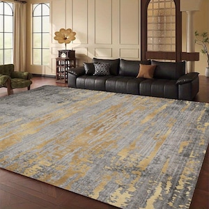 Puede incluir: Una alfombra rectangular grande con un diseño abstracto gris y dorado. La alfombra está colocada en un salón con un sofá de cuero negro y una mesa auxiliar de madera. El patrón de la alfombra tiene un efecto desgastado y pictórico.