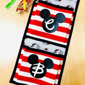 May include: A fabric wall hanging featuring two pockets with red and white horizontal stripes, each displaying a black Mickey Mouse silhouette with a white lowercase letter inside. The background fabric has a grey Mickey Mouse pattern.