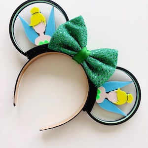 May include: A whimsical headband featuring black mouse ears with Tinkerbell designs. The ears have a clear frame with a silhouette of Tinkerbell, blue wings, and a yellow bun. A large, sparkly green bow sits between the ears.