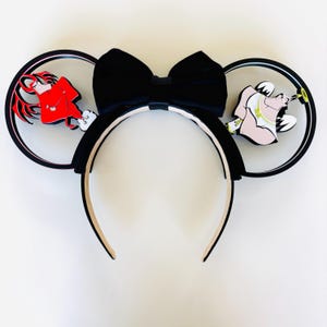 May include: A black headband featuring two circular ears with a large black bow in the center. The left ear displays a red devil character with wings, while the right ear features a white-clad angel character with a halo, both in a cartoon style.