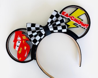 May include: Black headband with attached ears featuring a checkered bow, a red race car, and a yellow lightning bolt with the text "KA-CHOW!". The ears are black with a brown interior.