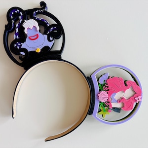 May include: A black headband with two Mickey Mouse ear-shaped decorations. One ear features Ursula the sea witch, and the other features Ariel the mermaid. The Ursula ear has a black frame with purple tentacles. The Ariel ear has a lavender frame with pink and green accents.