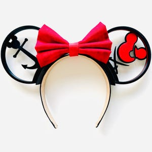 May include: A nautical-themed headband with black ears featuring an anchor and a red bow. The headband has a cream-colored interior and a black exterior. The bow is a vibrant red color, adding a pop of color to the design.
